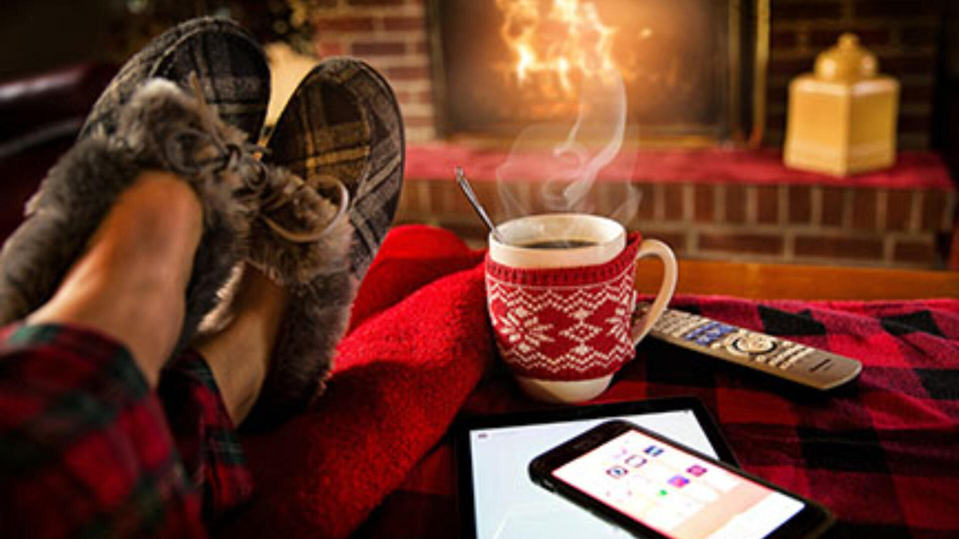 Eine Person in kariertem Pyjama und Hausschuhen entspannt sich vor einem Kamin und hat die Füße auf einer roten Decke hochgelegt. In der Nähe stehen eine Tasse mit Wintermuster, eine Fernbedienung, ein Tablet und ein Smartphone.